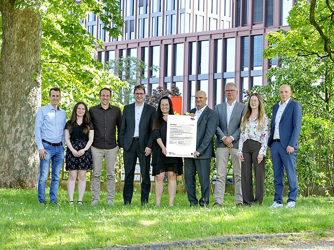 Gruppe von Menschen steht im Freien, eine Person hält ein Zertifikat, Gebäude im Hintergrund.