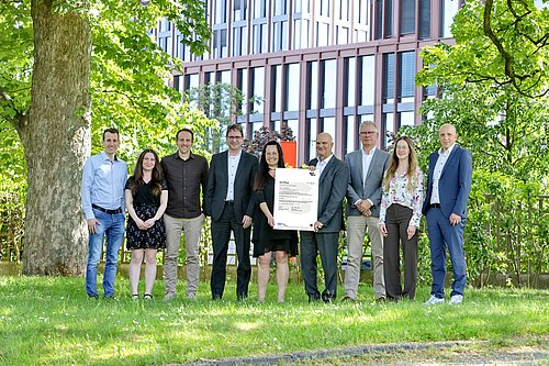 Gruppe von Menschen steht im Freien, eine Person hält ein Zertifikat, Gebäude im Hintergrund.