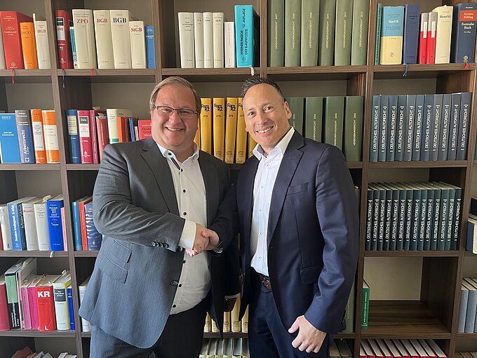Zwei Männer in Anzügen stehen lächelnd vor einem Bücherregal, das mit zahlreichen Fachbüchern gefüllt ist. Sie geben sich die Hand, was auf eine formelle Begrüßung oder ein Geschäftsabkommen hinweist.