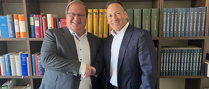 Zwei Männer in Anzügen stehen lächelnd vor einem Bücherregal, das mit zahlreichen Fachbüchern gefüllt ist. Sie geben sich die Hand, was auf eine formelle Begrüßung oder ein Geschäftsabkommen hinweist.
