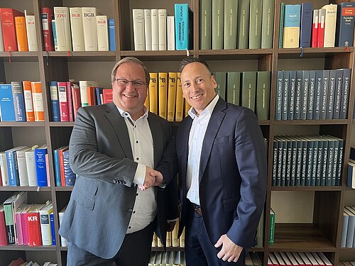 Zwei Männer in Anzügen stehen lächelnd vor einem Bücherregal, das mit zahlreichen Fachbüchern gefüllt ist. Sie geben sich die Hand, was auf eine formelle Begrüßung oder ein Geschäftsabkommen hinweist.