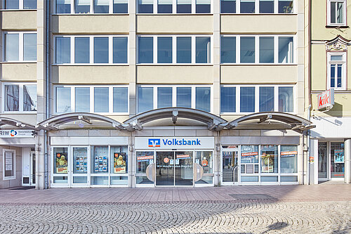 Eingang zur Volksbank-Filiale in einem mehrstöckigen Gebäude, mit großen Fenstern und Einkaufsmöglichkeiten daneben.