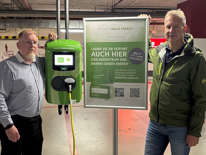Zwei Männer neben einer grünen BRAWO Green Energy Ladestation und einem Werbeplakat in einem Parkhaus.