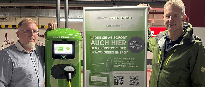 Zwei Männer neben einer grünen BRAWO Green Energy Ladestation und einem Werbeplakat in einem Parkhaus.