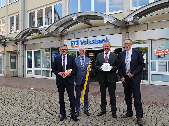 Vier Männer in Anzügen stehen vor einer Volksbank-Filiale. Einer hält Arbeitshandschuhe, ein anderer eine Wasserwaage, der nächste einen Bauhelm. Der Hintergrund zeigt den Gebäudeeingang mit dem Logo der Volksbank und einem Schild, das auf ein kostenloses Konto hinweist. Sie lächeln in die Kamera.