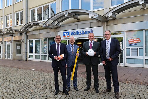 Vier Männer in Anzügen stehen vor einer Volksbank-Filiale. Einer hält Arbeitshandschuhe, ein anderer eine Wasserwaage, der nächste einen Bauhelm. Der Hintergrund zeigt den Gebäudeeingang mit dem Logo der Volksbank und einem Schild, das auf ein kostenloses Konto hinweist. Sie lächeln in die Kamera.